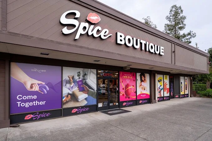 The Spice Boutique storefront in Santa Rosa — pink illuminated 'Spice BOUTIQUE' signage above the door, bright OPEN sign, large window displays facing a clean storefront walkway with mature trees behind the building